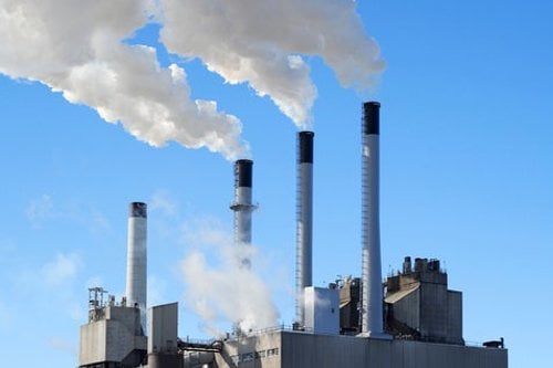 Industrial power plant with four smokestacks emitting white smoke against a clear blue sky