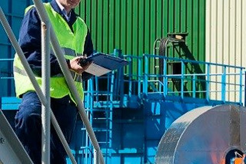 Kiwa auditor in safety gear performing an inspection with a clipboard at a manufacturing facility, featuring machinery and industrial equipment in the background