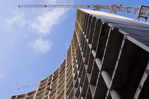 Bouwconstructie van een hoog gebouw met twee bouwkranen op de achtergrond tegen een blauwe lucht.