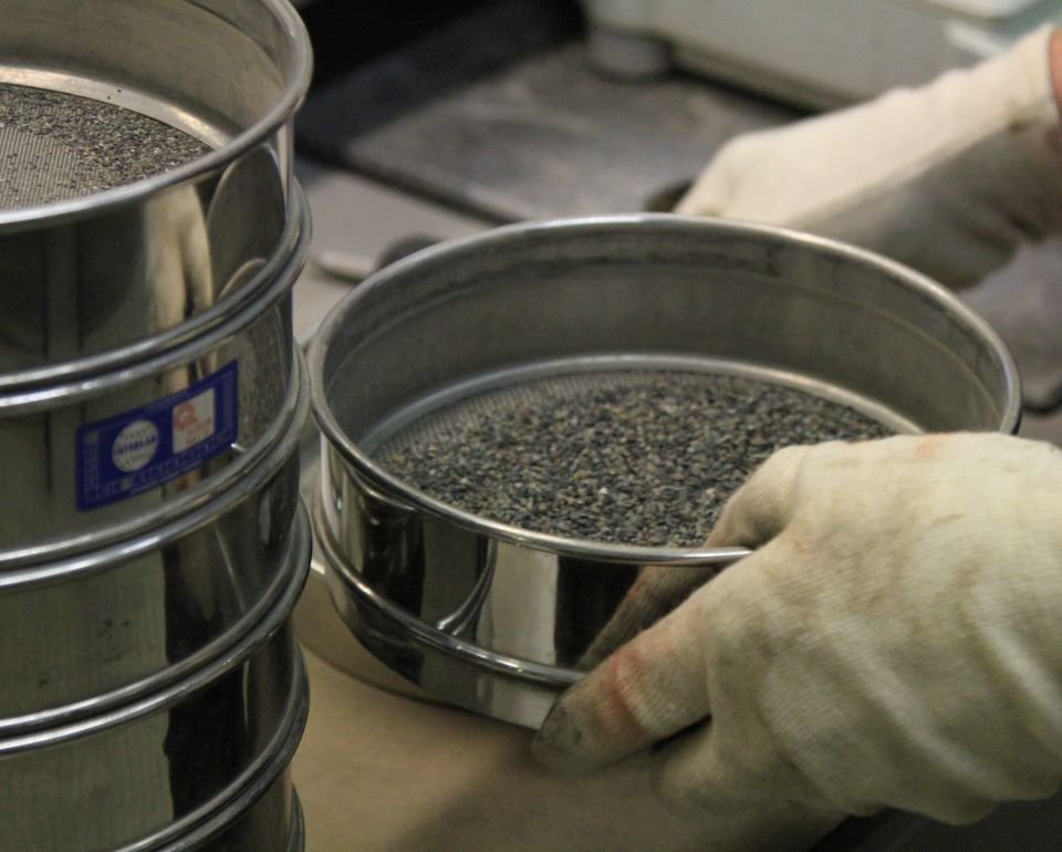 Handen met handschoenen controleren zeef met losgekomen asfalt in een laboratoriumomgeving.