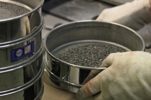 Handen met handschoenen controleren zeef met losgekomen asfalt in een laboratoriumomgeving.
