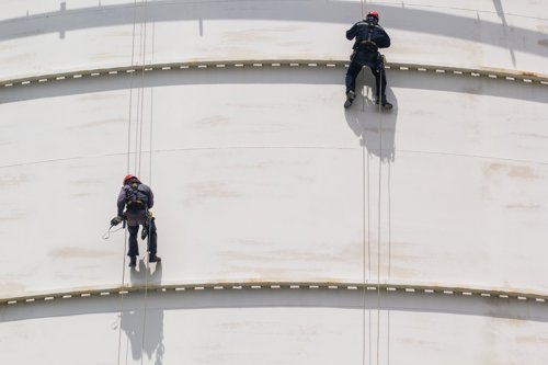 Twee industriële klimmers met veiligheidsuitrusting hangen aan touwen tegen een grote witte metalen tank.