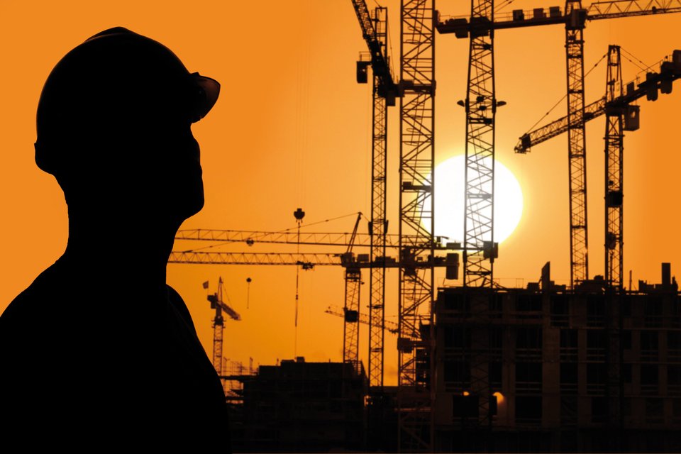 Silhouet van een persoon met een bouwhelm voor een achtergrond van hijskranen en gebouwen bij zonsondergang.