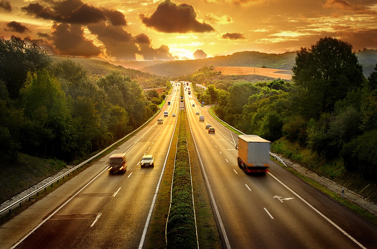 Snelweg met meerdere rijstroken omgeven door groene bomen en heuvels bij zonsondergang.