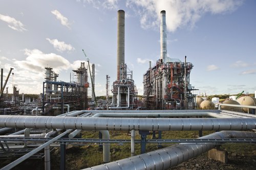 Industrieel energiecomplex met grote pijpleidingen, metalen installaties en twee hoge schoorstenen onder een deels bewolkte hemel.