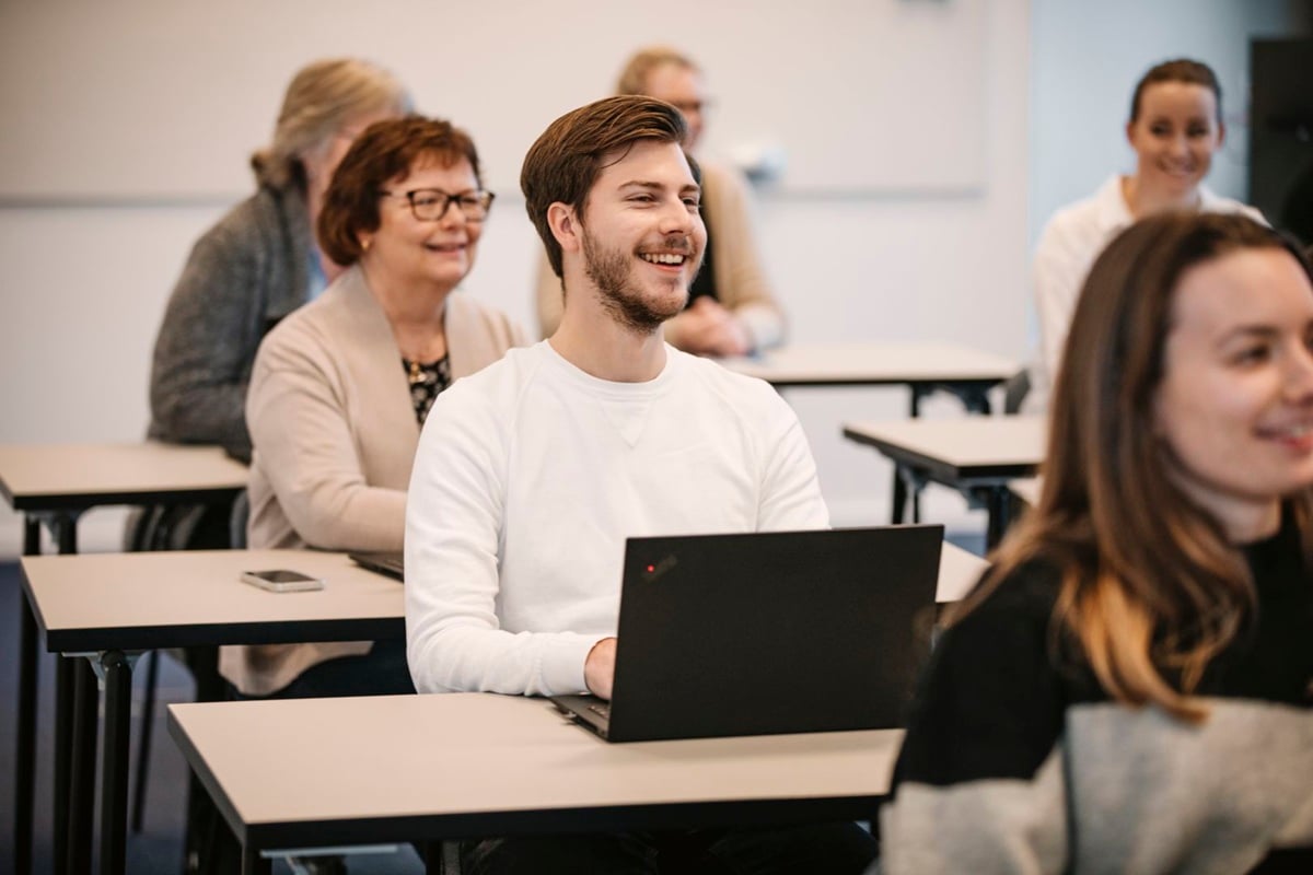 Groep volwassenen in een klaslokaal met laptops aan tafels tijdens een training of vergadering.
