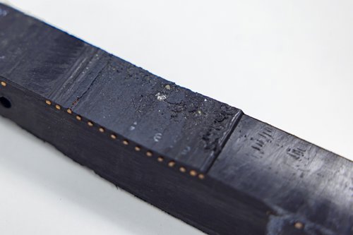 Zwart rechthoekig object met een matte afwerking, zichtbaar slijtage en kleine details op het oppervlak, op een witte achtergrond.