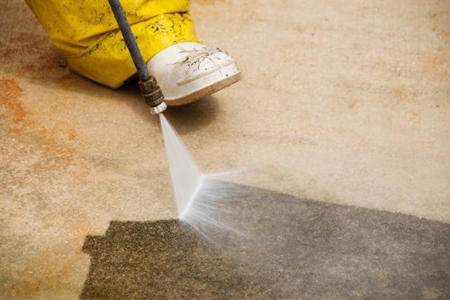 Een persoon met gele laarzen gebruikt een hogedrukreiniger om een vuil oppervlak schoon te maken.
