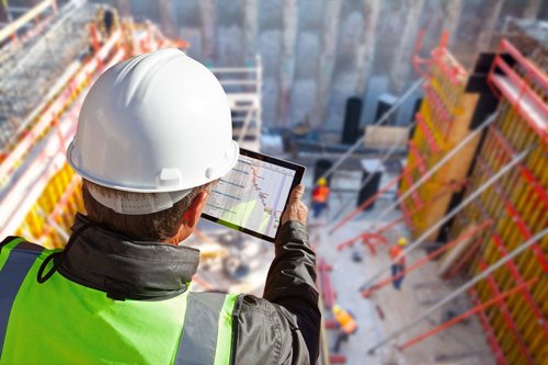 Man met witte helm en fluorescerend veiligheidsvest bekijkt bouwtekeningen op een tablet op een bouwplaats.