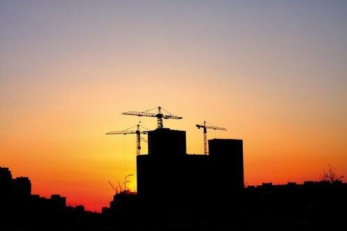 Silhouet van gebouwen in aanbouw met kraanmachines tegen een kleurrijke zonsonderganghemel.