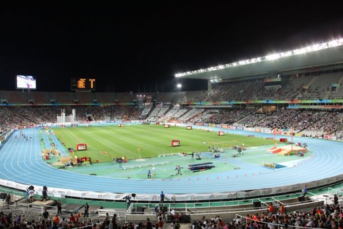 Atletiekstadion bij nacht, gevuld met toeschouwers, een blauwe atletiekbaan en verschillende sportarena's op het veld.