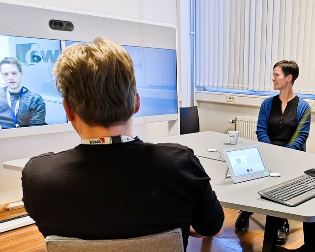 Cilvēki sēž pie galda konferenču telpā, ar ekrānu, uz kura redzami attēli, un datoriem priekšplānā.