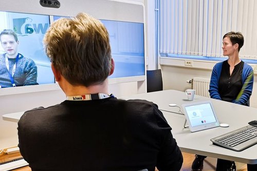 Cilvēki sēž pie galda konferenču telpā, ar ekrānu, uz kura redzami attēli, un datoriem priekšplānā.