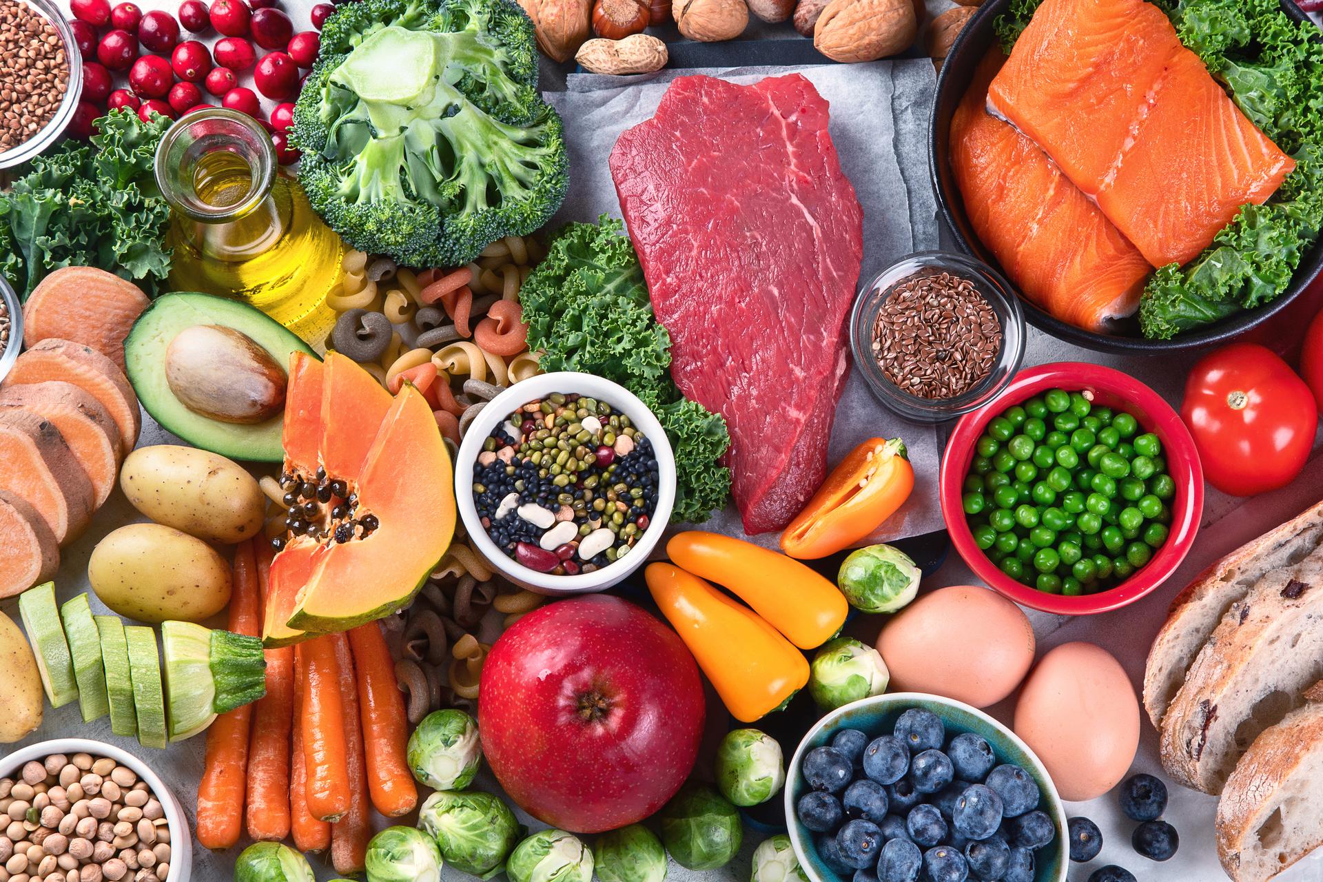 Assortment of fresh healthy foods including salmon, beef, vegetables, fruit, eggs, beans, bread, and whole grains arranged on a table.