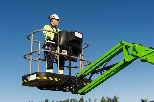 Arbetare i säkerhetsutrustning står i en lyftplattform på en grön bomlift mot klarblå himmel - Kiwa erbjuder en kurs för mobila arbetsplattformer för att förebygga farliga situationer.