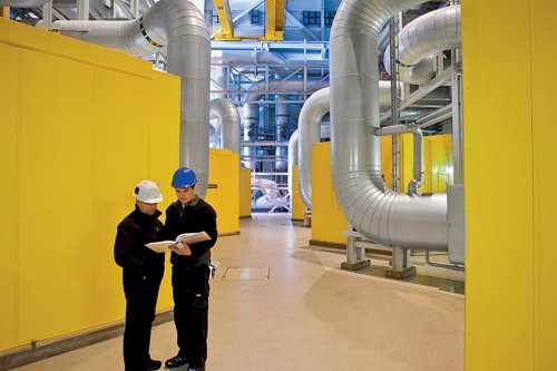 Två personer i arbetskläder och hjälmar står och håller dokument i en industriell anläggning med stora rör och trycksatta anordningar med gula väggar - Kiwa erbjuder en kurs i arbetet med att skapa livslängdsjournaler och skademekanismer.