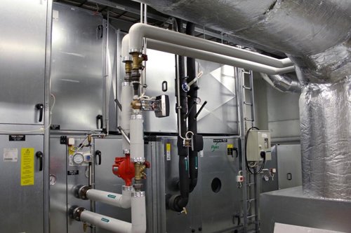 Tekniskt ventilationssystem i maskinrum med isolerade ventilationskanaler, rör, ventiler och stora metallskåp i en bild som förmedlar behovet av certifierade injusterare av ventilationssystem.