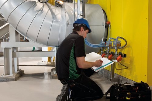 Tekniker med blå hjälm som knäböjer och utför Fitness for Service vid industriell rörinstallation med stora silverfärgade ventilationskanaler och gul vägg.