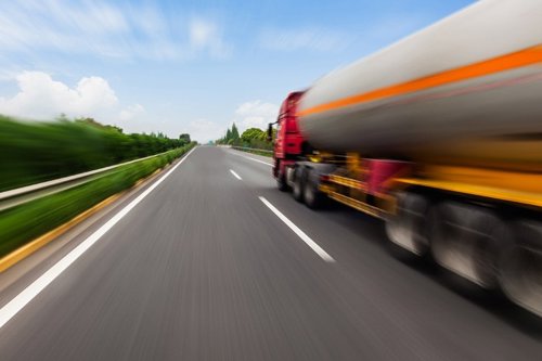 Tung lastbil med släpvagn kör snabbt på en motorväg omgiven av gröna träd under en klarblå himmel.