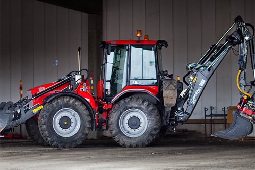 Röd traktor med grävutrustning, parkerad inomhus på en verkstadsgolv.