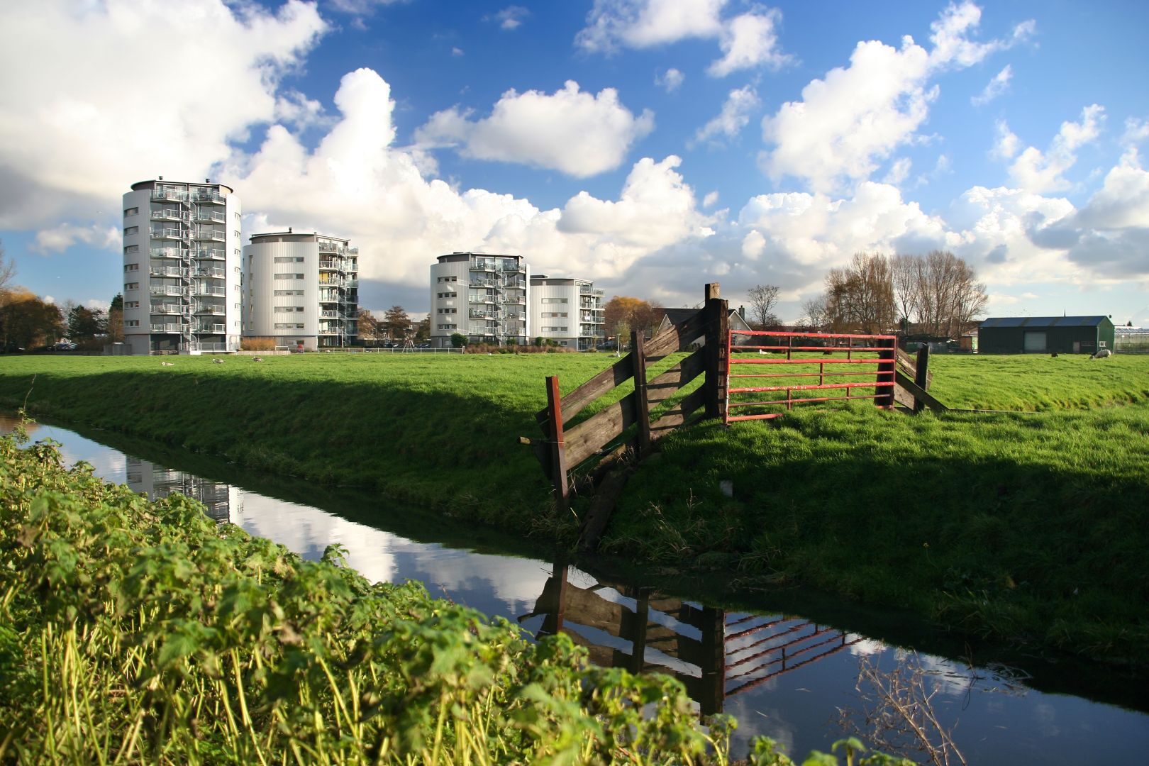 Ett gräsbevuxet landskap vid floden med moderna flerbostadshus, en röd grind och en kanal under en molnig blå himmel.