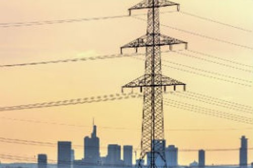 Augstsprieguma elektrolīnija lauku ainavā ar pilsētas panorāmu fonā, saulrieta gaismā.