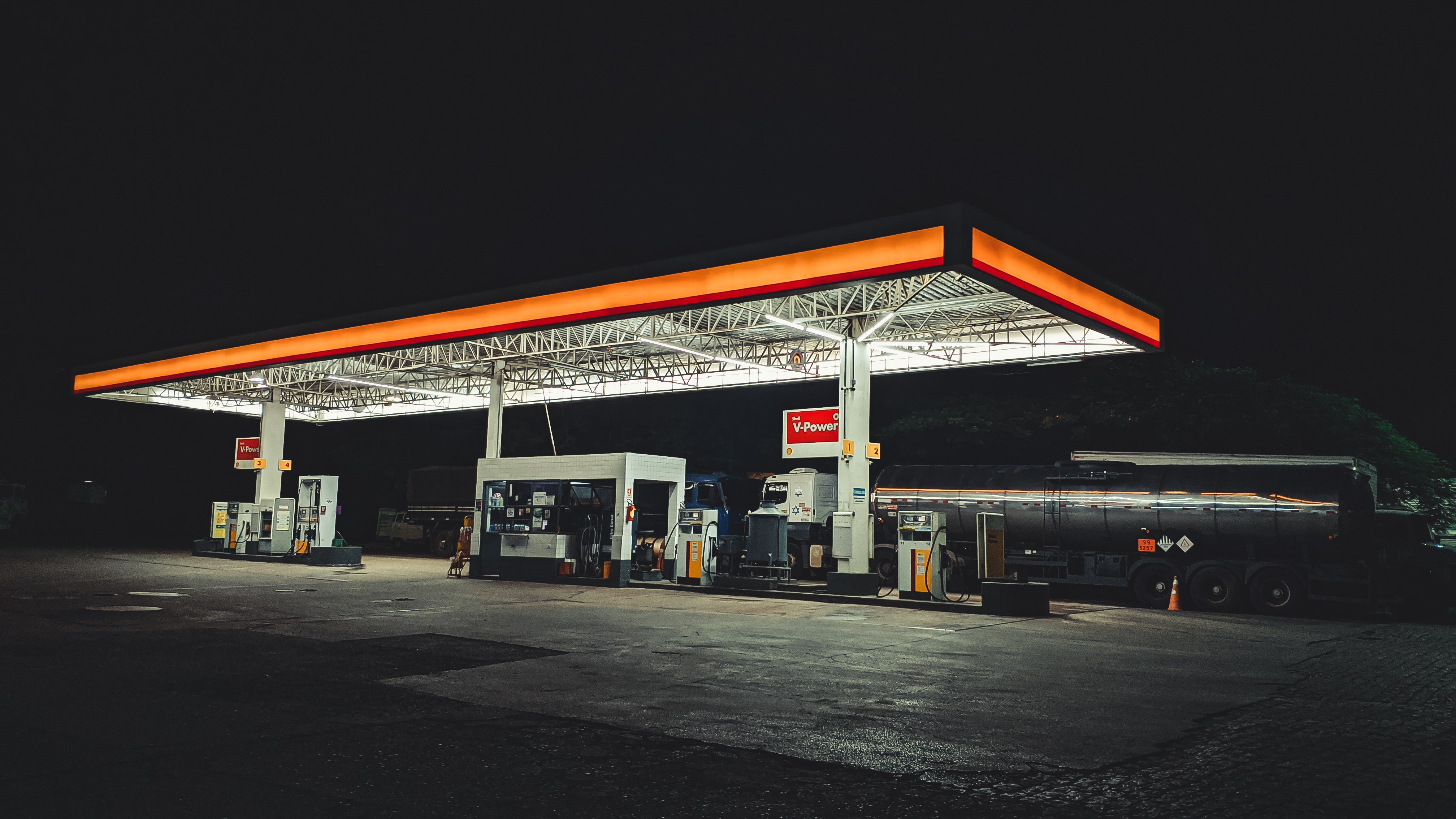 Degvielas uzpildes stacija naktī ar apgaismotu nojumi, uzpildes sūkņiem un cisternas kravas automašīnu.