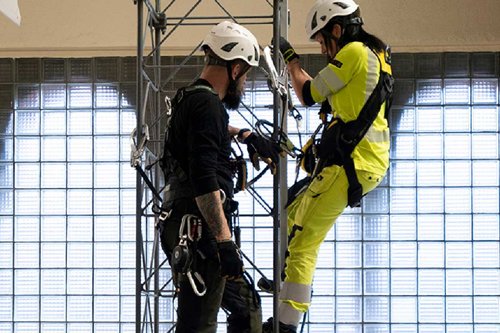 Två personer med säkerhetsutrustning klättrar på en metallstruktur, iklädda skyddshjälmar och arbetskläder.