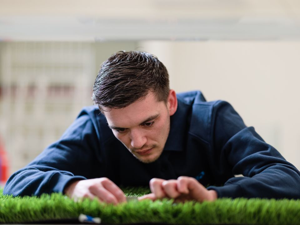 Man liggend voor een model van kunstgras, gekleed in een donkerblauw shirt, met focus op de grasstructuur.