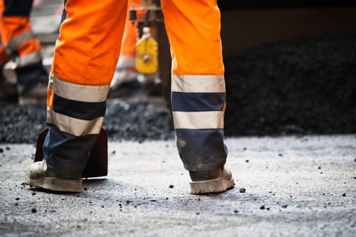 Persoon in oranje werkbroek met reflecterende strepen bij een wegwerkzaamheden, omringd door asfalt en gereedschap.