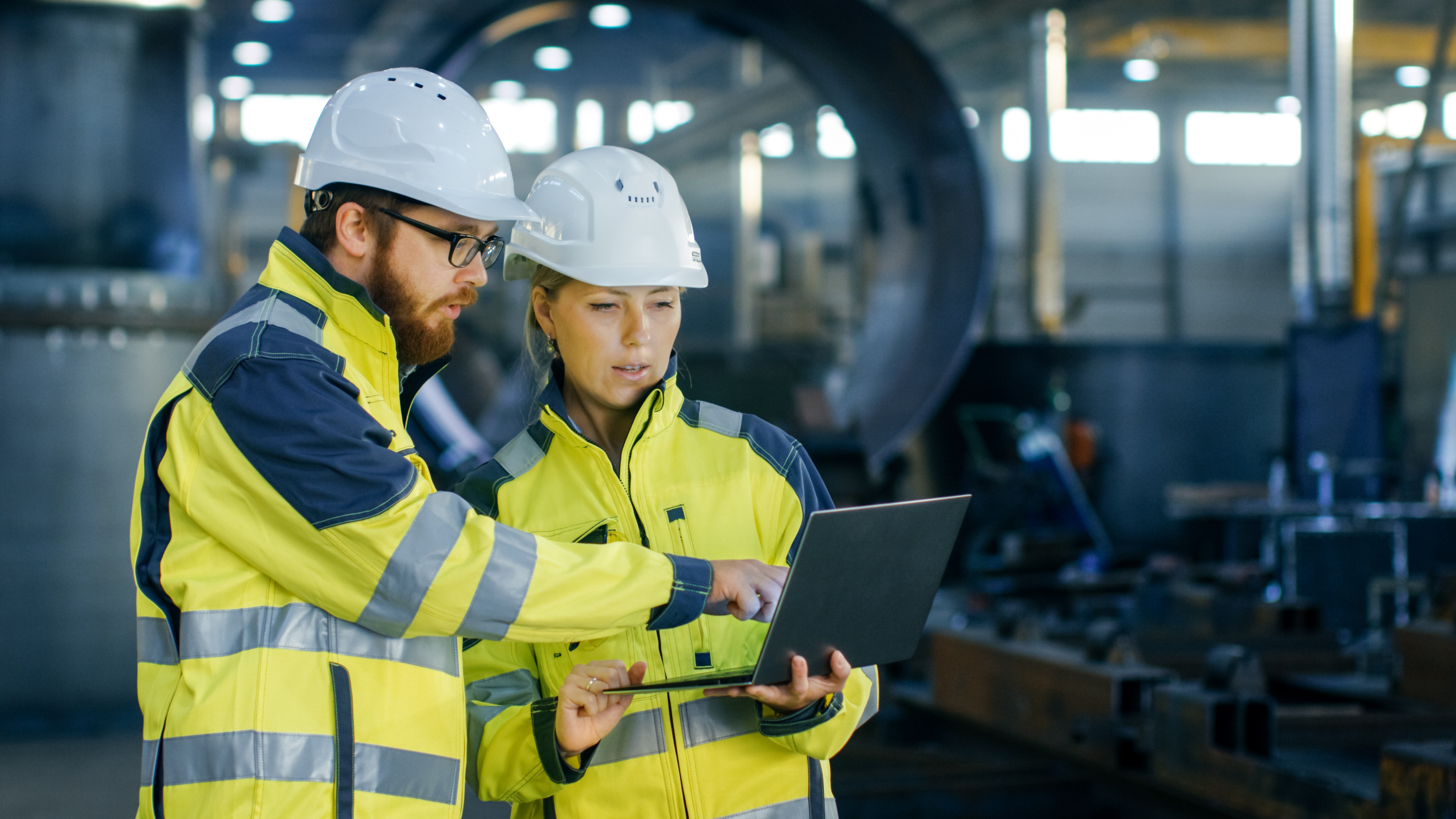 Twee inspecteurs in gele veiligheidskleding en witte helmen overleggen met een laptop in een industriële werkomgeving