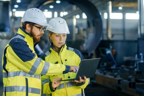 Twee inspecteurs in gele veiligheidskleding en witte helmen overleggen met een laptop in een industriële werkomgeving