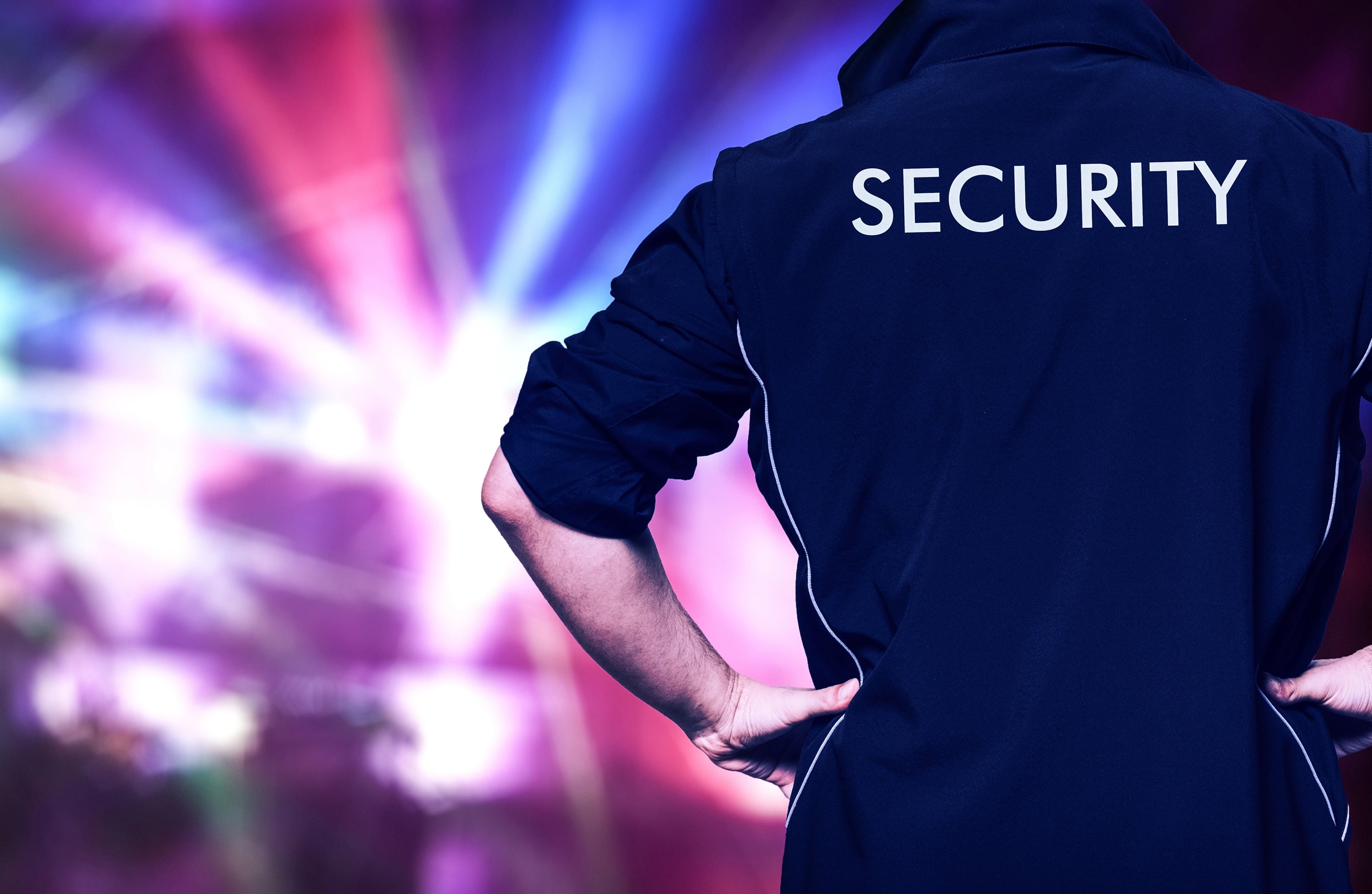 Man in een donkerblauw jack met de tekst "SECURITY" op de rug, staande voor een kleurrijke achtergrond met lichteffecten.