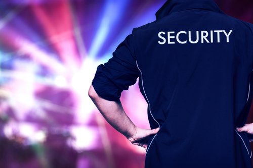 Man in een donkerblauw jack met de tekst "SECURITY" op de rug, staande voor een kleurrijke achtergrond met lichteffecten.