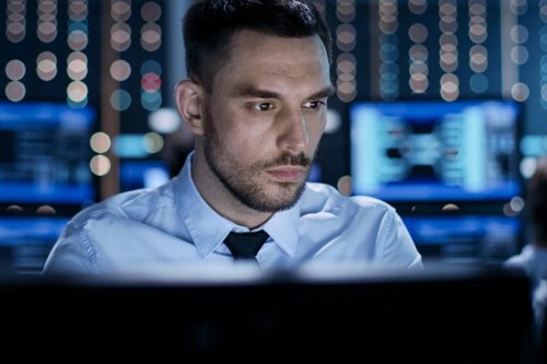 Man zittend aan een bureau in een moderne controlekamer, met achtergrond van computerschermen en LED-verlichting.