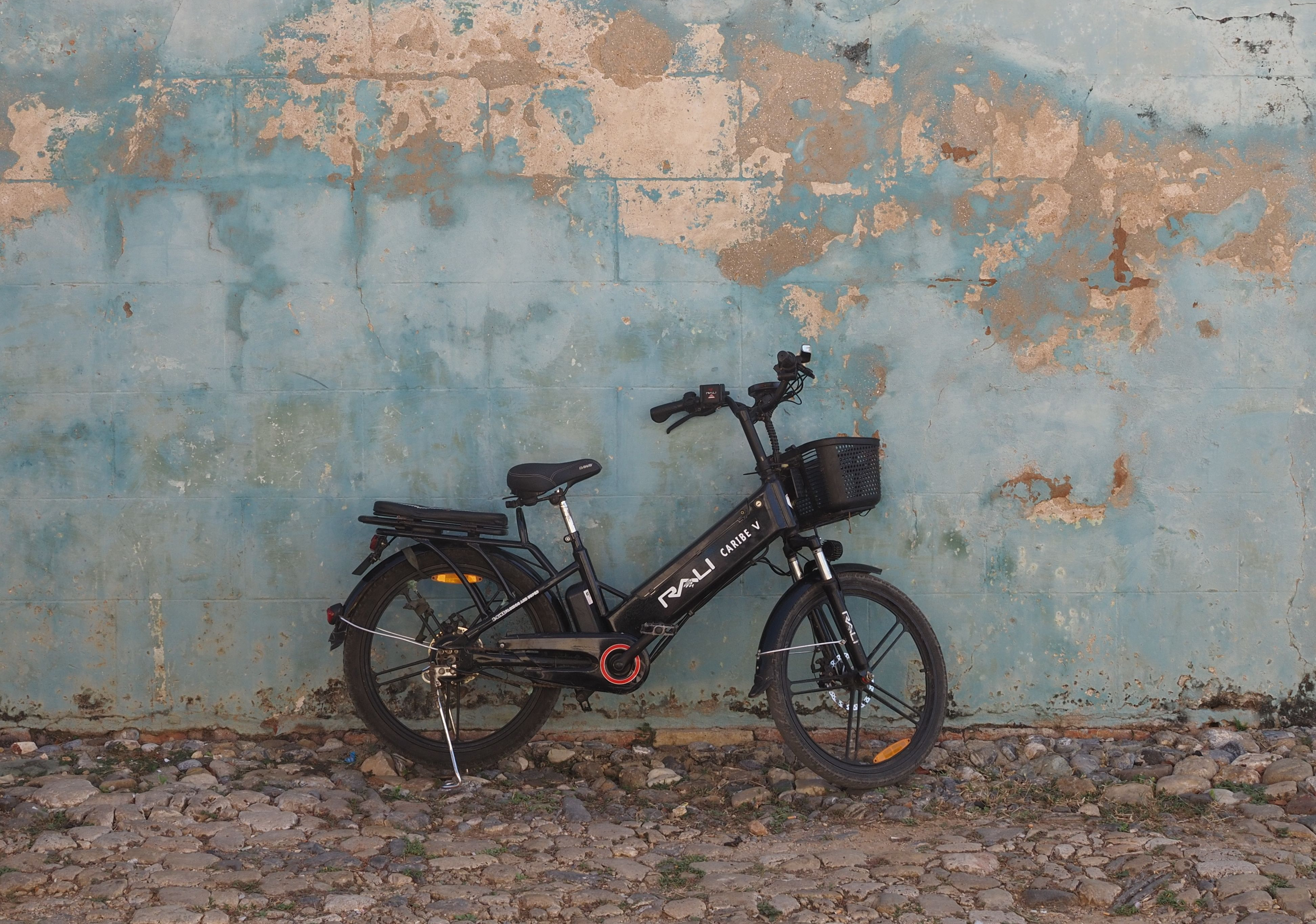 Zwarte elektrische fiets met mand aan het stuur, geparkeerd tegen een verweerde blauwgroene muur met afbladderende verf. De fiets staat op een ondergrond van keien en grind, met een minimalistische en rustige achtergrond zonder andere objecten.