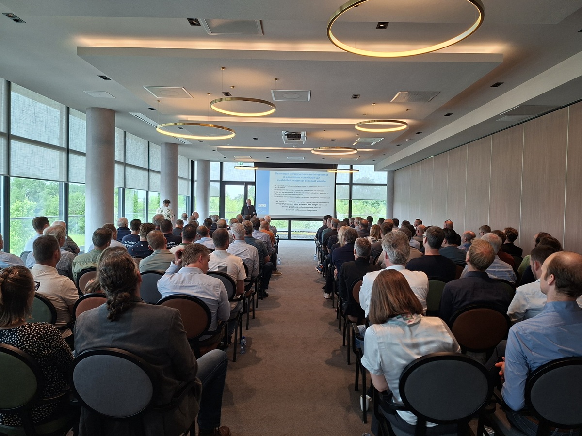 Zakelijke conferentie in een moderne vergaderzaal met een publiek dat naar een presentatie op een projectiescherm kijkt. Grote ramen, ronde plafondlampen en rijen stoelen vullen de ruimte.