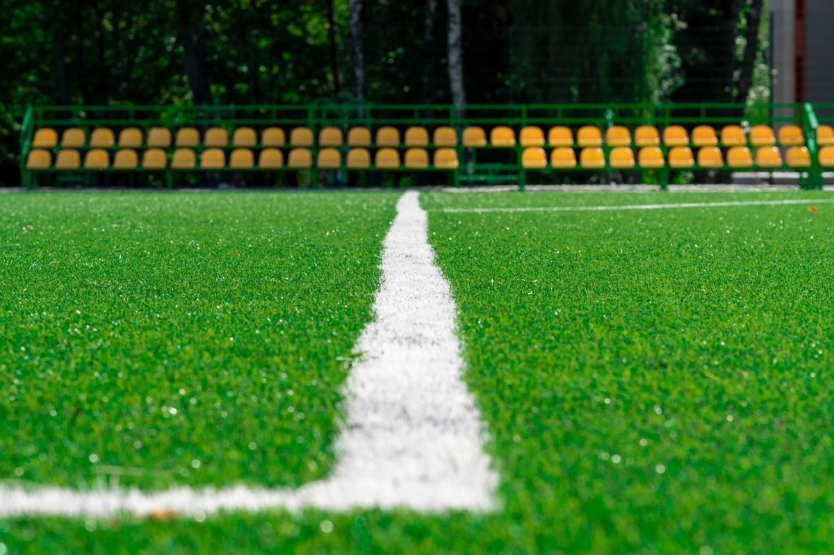 Close-up van een groen kunstgras sportveld met witte veldlijn en gele stadionstoelen op de achtergrond.