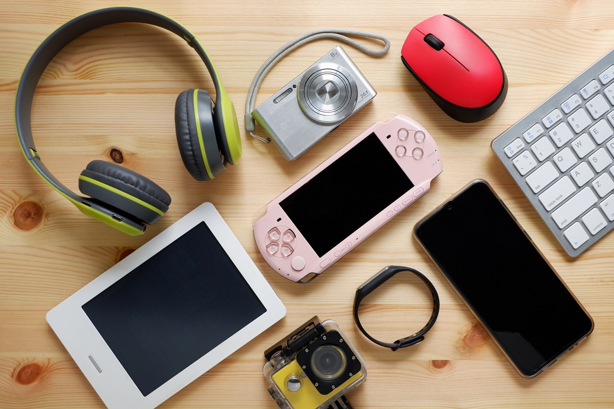 Top view of assorted electronics on a wooden table, including headphones, cameras, a handheld game console, a tablet, smartphones, a keyboard, a mouse, and a wristband.
