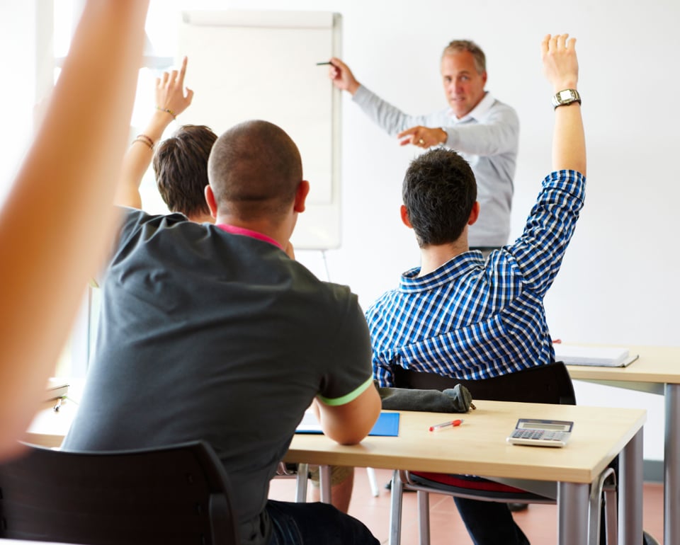 Students raising hands in a classroom while a Kiwa instructor stands in front of a whiteboard, engaged in a discussion.
