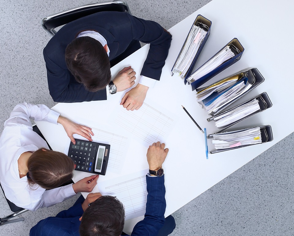 Overhead view of three office workers reviewing documents and using a calculator at a white conference table with organized binders and paperwork.