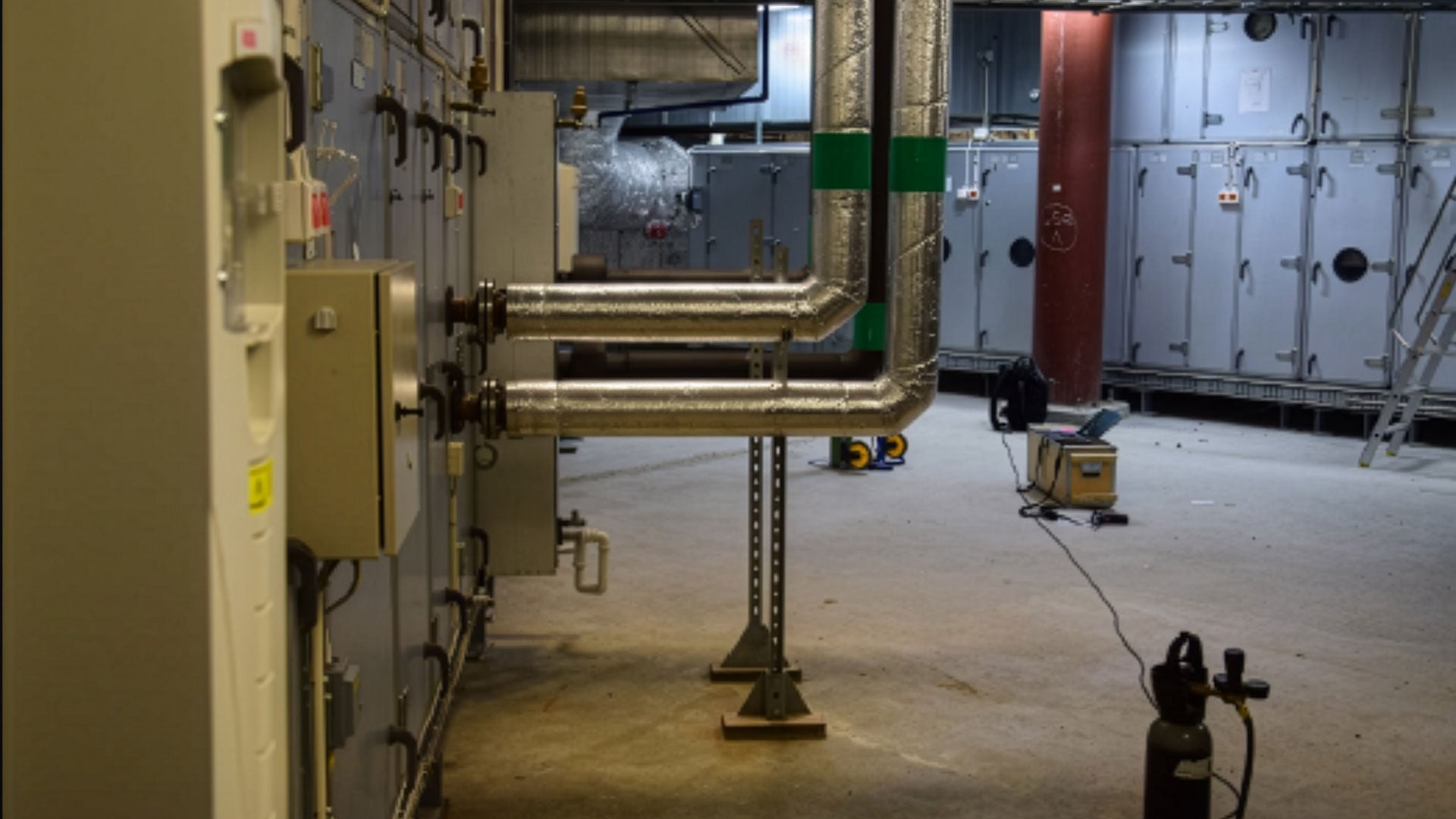 Spårgasmätning pågår i ett ventilationsrum med stora rör, isolerade kanaler och industriella aggregat i en byggnad.