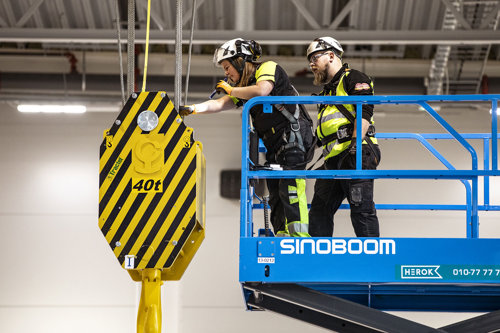 Två besiktningsingenjörer arbetar på en blå arbetsplattform och inspekterar en stor gul och svart lyftkrok märkt med "40t" i en industriell miljö.