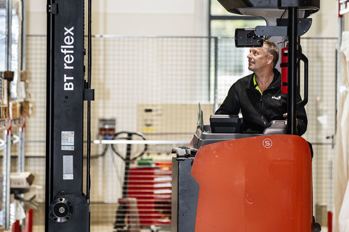Besiktningsingenjör kontrollerar och kör en orange BT Reflex-styvtruck i en lagerbyggnad med hyllor och utrustning i bakgrunden.