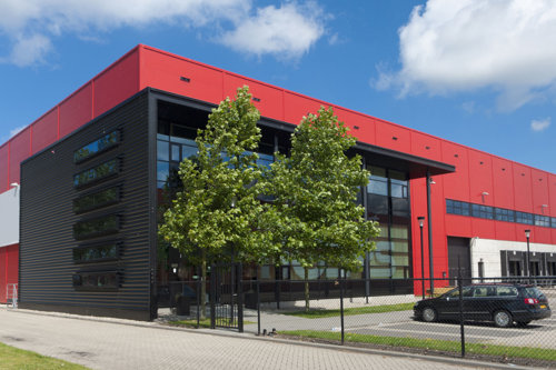 Stor röd och svart industribyggnad med stora fönster, träd framför entrén.