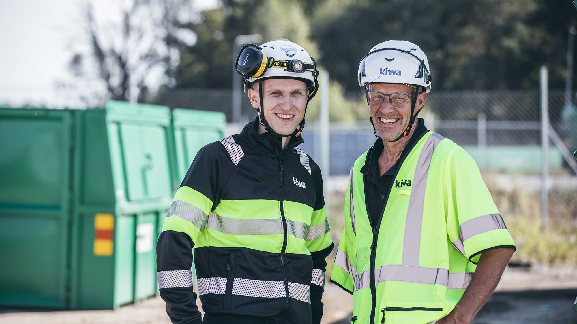Två personer i skyddskläder och hjälmar står utomhus på en byggarbetsplats bredvid en grön container.