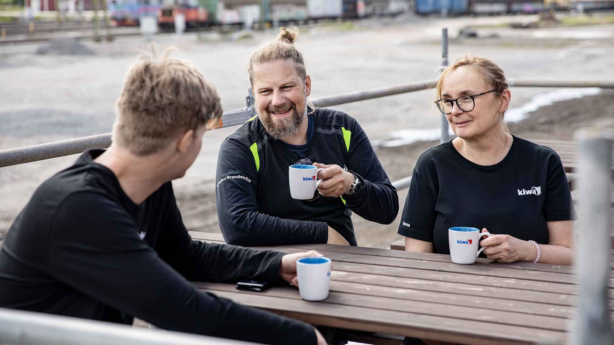 Tre personer i svarta tröjor sitter vid ett träbord utomhus och dricker kaffe ur vita muggar.