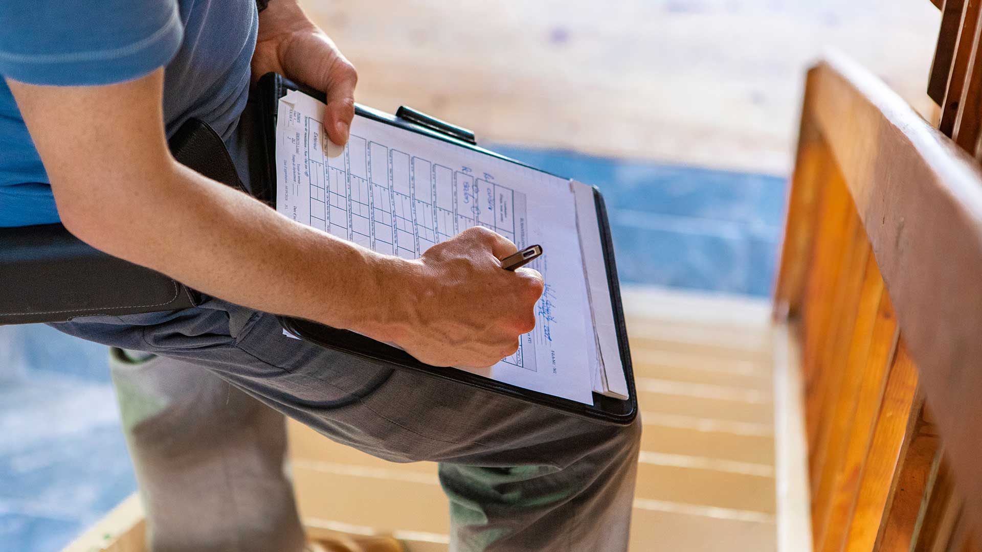 Överlåtelsebesiktningstekniker fyller i ett formulär på ett clipboard med penna, nära en trappa inomhus.