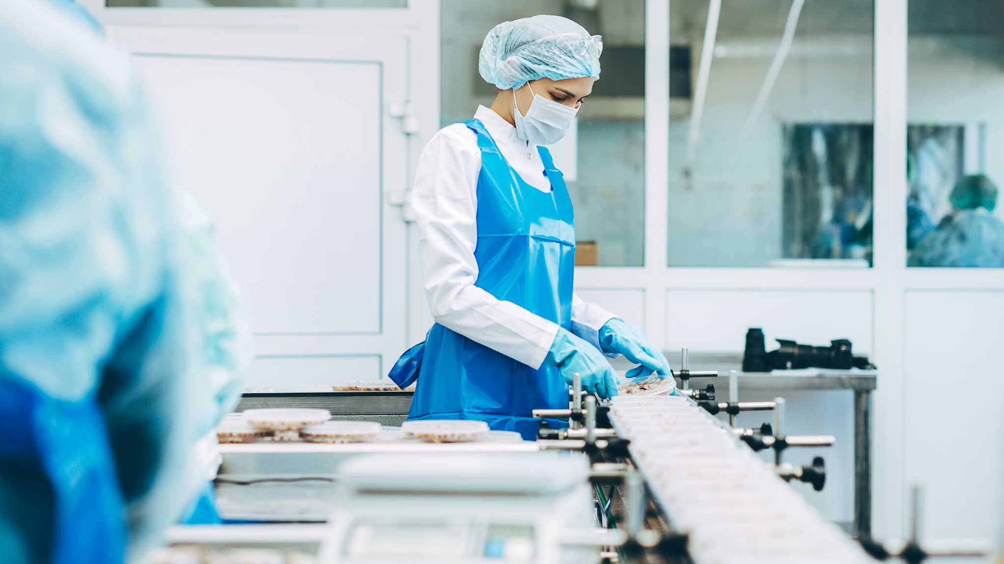 Person i skyddskläder och hårnät som arbetar vid ett produktionsband i en livsmedelsfabrik.