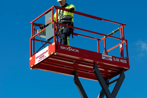 En person står i en röd skylift mot en klarblå himmel, iförd gul skyddsklädsel och säkerhetsutrustning.